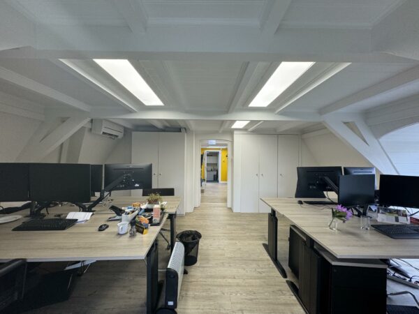 Modern kantoor met houten vloer, meerdere bureaus met computers en zicht op een gang met gele muur achterin.